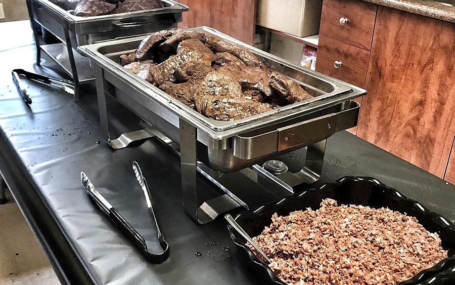 A catering spread: chafing dishes of braised beef with serving tongs, a tray of bacon bits and chopped green onions